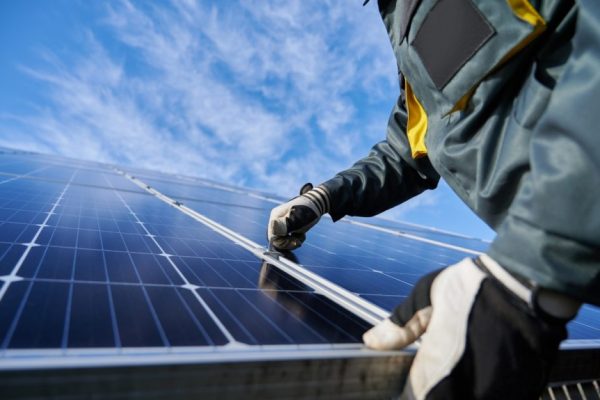 Close up of man technician in work gloves installing stand-alone photovoltaic solar panel system under beautiful blue sky with clouds. Concept of alternative energy and power sustainable resources.
